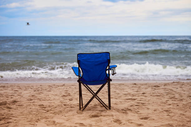 Blue folding chair back on sea beach, without people, beach holiday alone, loneliness Blue folding chair back on seashore, sea background. Blue chair on sea beach, without people. Beach holiday alone, nobody on Baltic Sea beach, loneliness and hopelessness folding chair stock pictures, royalty-free photos & images