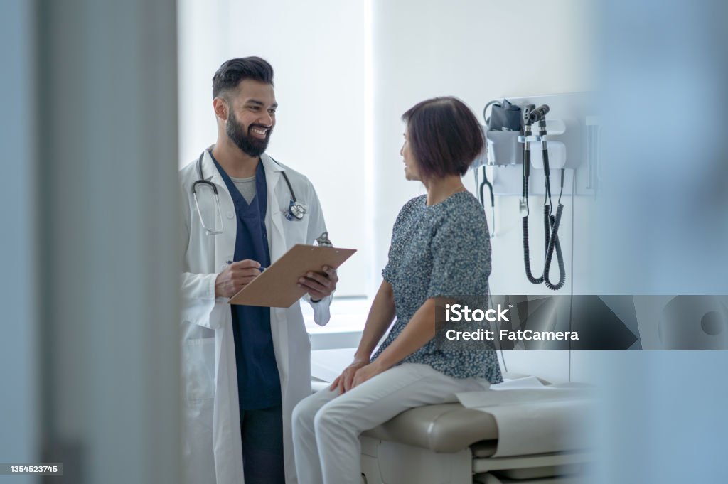 Senior Talking with the Doctor About a Flu Shot A senior woman sit up on the exam table as she talks with her male doctor about the benefits of the Flu Shot.  She is dressed casually and listening attentively as her doctor holds out a clipboard of information and they discuss it. Doctor Stock Photo