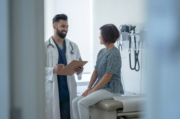 Senior Talking with the Doctor About a Flu Shot A senior woman sit up on the exam table as she talks with her male doctor about the benefits of the Flu Shot.  She is dressed casually and listening attentively as her doctor holds out a clipboard of information and they discuss it. doctor consultation stock pictures, royalty-free photos & images