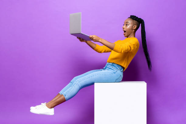 mujer afroamericana emocionada sosteniendo y mirando la computadora portátil mientras está sentada en el taburete en un fondo de estudio de color púrpura aislado - red chairs fotografías e imágenes de stock