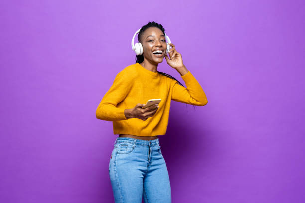 African American woman wearing headphones listening to music from smart phone on colorful purple isolated studio background stock photo