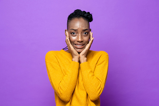 Young African-American woman feeling fear and frightened with hands on cheeks in purple isolated studio background