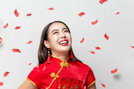 Smiling happy Asian woman in traditional oriental costume looking upward in light gray background with red confetti
