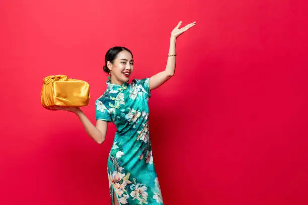 Smiling pretty Asian woman in oriental style costume with golden gift box in red isolated studio background for Chinese new year concepts