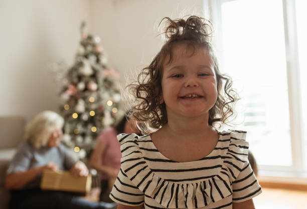 A cute little girl is smiling, standing in the middle of the living room. Behind her are her mom, granny, and a big beautiful Christmas tree. One little girl is standing in the center of the living room.  She is smiling and enjoying during the Winter holidays with her family. Behind her are granny and mom, with New Year's presents in their hands. baby new years eve new years day new year stock pictures, royalty-free photos & images