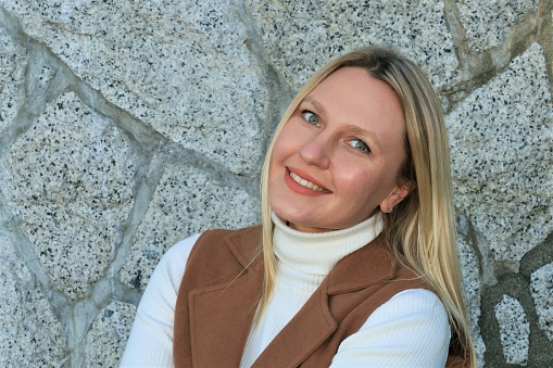 A blond woman with healthy teeth and great smile. She is wearing a white sweater and brown sleeveless coat.