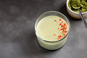 One matcha panna cotta dessert with confectionery red decor in a glass with spoon on grey background. Matcha tea next to it.