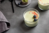 Matcha panna cotta dessert with confectionery red decor with blueberry in a glass with spoon on grey background. Matcha tea next to it. Minimalistic photography.