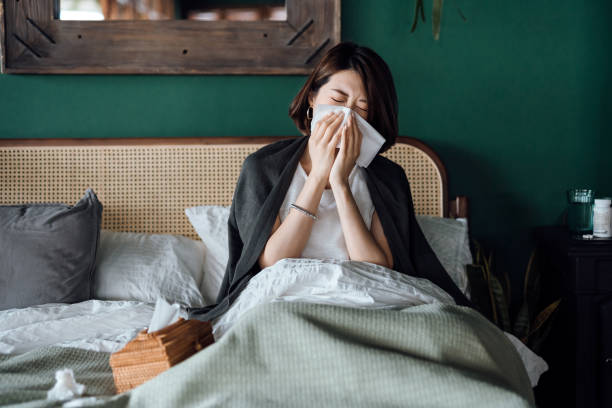 giovane donna asiatica seduta sul letto e soffiarsi il naso con un fazzoletto mentre soffre di raffreddore, con bottiglia di medicina e un bicchiere d'acqua sul tavolino - cold and flu flu virus sneezing illness foto e immagini stock