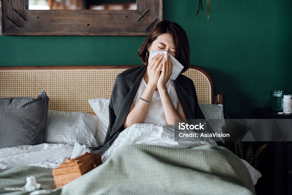 Young Asian woman sitting on bed and blowing her nose with tissue while suffering from a cold, with medicine bottle and a glass of water on the side table Cold And Flu Stock Photo