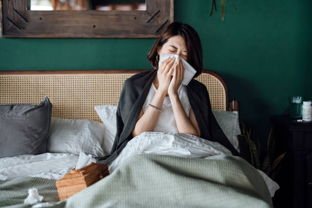 Young Asian woman sitting on bed and blowing her nose with tissue while suffering from a cold, with medicine bottle and a glass of water on the side table stock photo