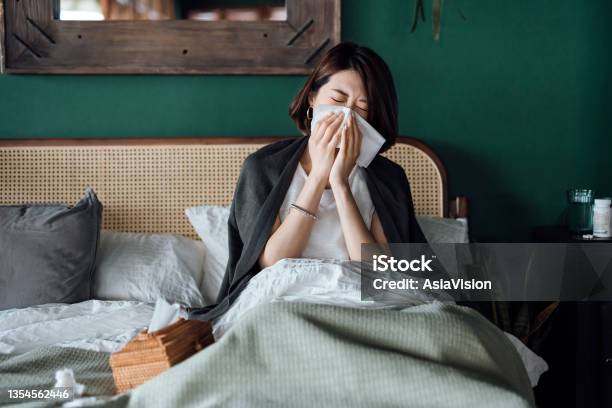 Young Asian Woman Sitting On Bed And Blowing Her Nose With Tissue While Suffering From A Cold With Medicine Bottle And A Glass Of Water On The Side Table Stock Photo - Download Image Now