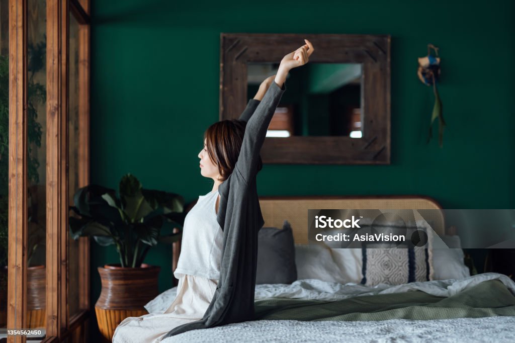 Side profile of young Asian woman sitting in bed, stretching arms after waking up in the morning against sunlight. Let's get the day started Waking up Stock Photo