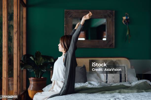 Side Profile Of Young Asian Woman Sitting In Bed Stretching Arms After Waking Up In The Morning Against Sunlight Lets Get The Day Started Stock Photo - Download Image Now