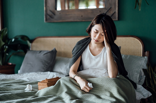 Young Asian woman feeling sick and suffering from a headache, massaging forehand to relieve the pain, sitting on the bed and taking a rest at home