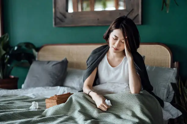 Photo of Young Asian woman feeling sick and suffering from a headache, massaging forehand to relieve the pain, sitting on the bed and taking a rest at home