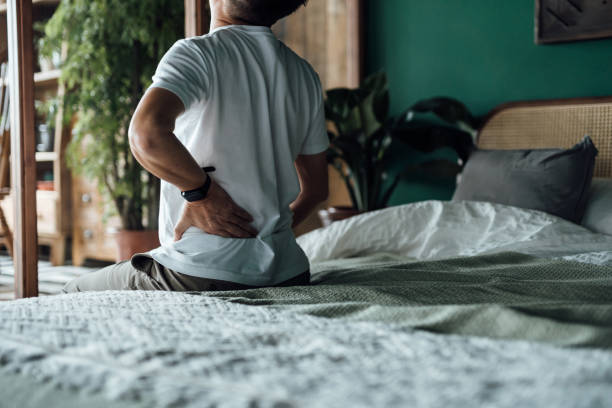 vista posteriore dell'uomo asiatico anziano che soffre di mal di schiena, massaggiando i muscoli doloranti mentre è seduto sul letto. concetto di anziani e problemi di salute - schiena foto e immagini stock