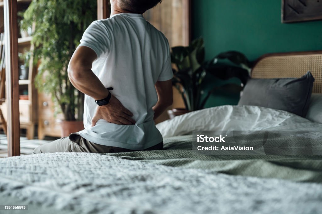 Rear view of senior Asian man suffering from backache, massaging aching muscles while sitting on bed. Elderly and health issues concept Backache Stock Photo