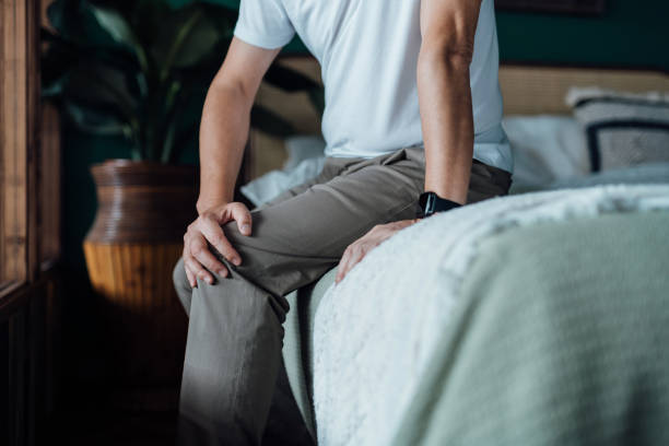 Close up of senior man holding his knee in discomfort, suffering from knee pain while sitting on bed at home. Elderly and health issues concept stock photo
