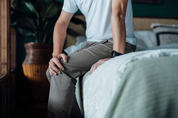 close up of senior man holding his knee in discomfort, suffering from knee pain while sitting on bed at home. elderly and health issues concept - legs only imagens e fotografias de stock