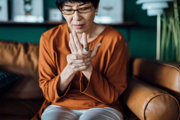 Senior Asian woman rubbing her hands in discomfort, suffering from arthritis in her hand while sitting on sofa at home. Elderly and health issues concept stock photo