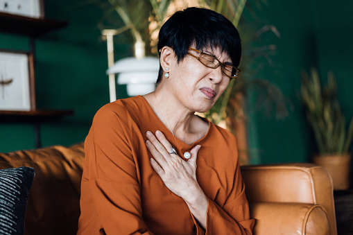 Senior Asian woman with eyes closed holding her chest in discomfort, suffering from chest pain while sitting on sofa at home. Elderly and health issues concept