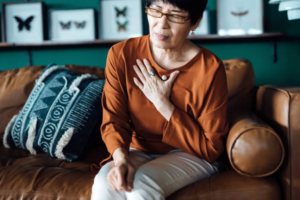senior asian woman with eyes closed holding her chest in discomfort, suffering from chest pain while sitting on sofa at home. elderly and health issues concept - indigestie fotos stockfoto's en -beelden