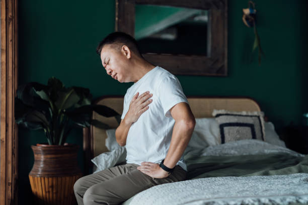 Senior Asian man with eyes closed holding his chest in discomfort, suffering from chest pain while sitting on bed at home. Elderly and health issues concept stock photo
