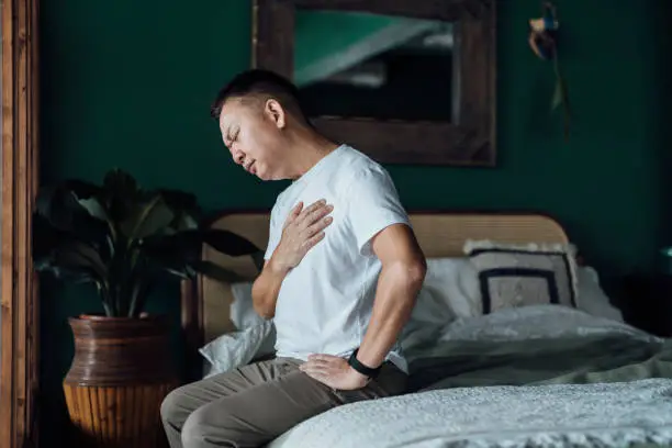 Photo of Senior Asian man with eyes closed holding his chest in discomfort, suffering from chest pain while sitting on bed at home. Elderly and health issues concept