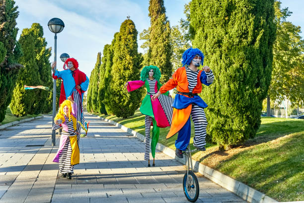 Full body troupe of funny creative performers in colorful harlequins costumes and wigs, walking on stilts and riding unicycle during parade on sunny day in park