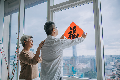 Chinese new year senior couple decorating house living room with chinese calligraphy decoration on window preparing for family reunion