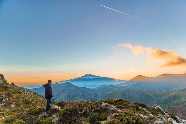 man standing on top of the cliff in the mountains at sunset enjoying the beautiful sunset over erupting volcano etna. beautiful view in sicily, italy - mt etna imagens e fotografias de stock