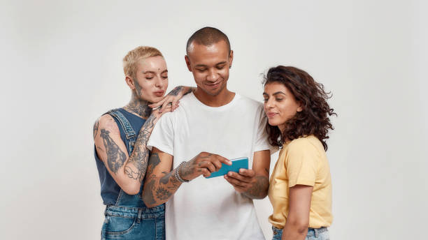 スマートフォンを使ったカジュアルウェア、ビデオを見て、光の背景に孤立した一緒に立っている3人の若い多様な人々 - men young adult human head blond hair ストックフォトと画像