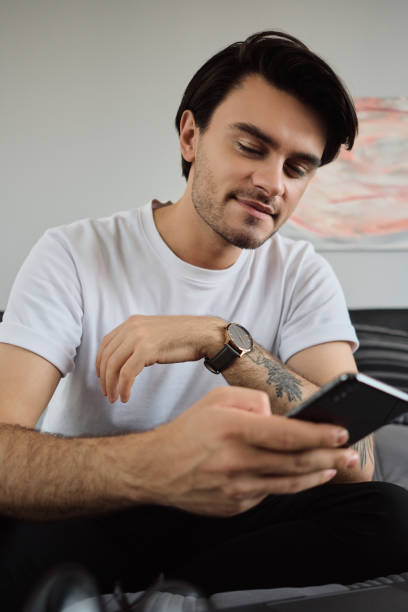 istock Young handsome smiling brunette man in white T-shirt dreamily using cellphone sitting on bed with laptop at modern home 1354601207