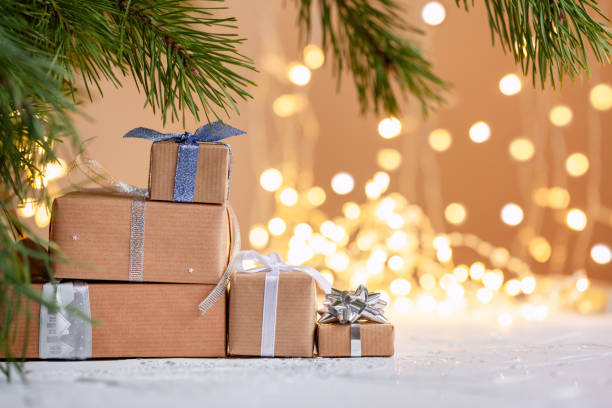 Stacks of Christmas gift boxes laid under a Christmas tree with defocused lights. Handmade surprise presents. New year holiday concept.