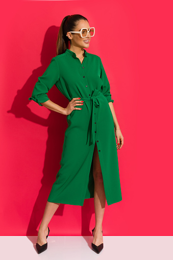 Beautiful woman in long button-up green dress, sunglasses and high heels is posing with hand on hip, and looking away. Full length on red background.