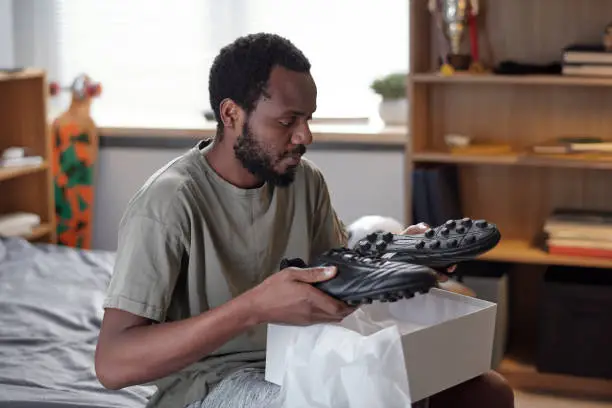 African man holding pair of new black trendy sports-shoes over while open box while unpacking sports footwear