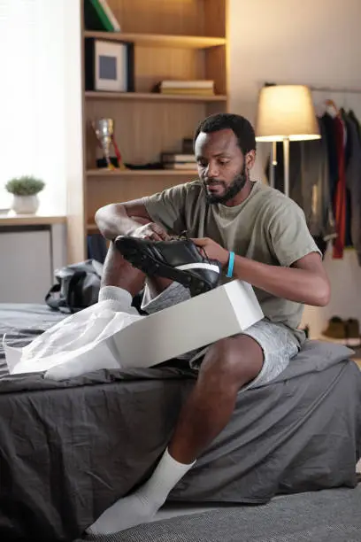 Young African sportsman in activewear unpacking box with new black sports-shoes while sitting on bed