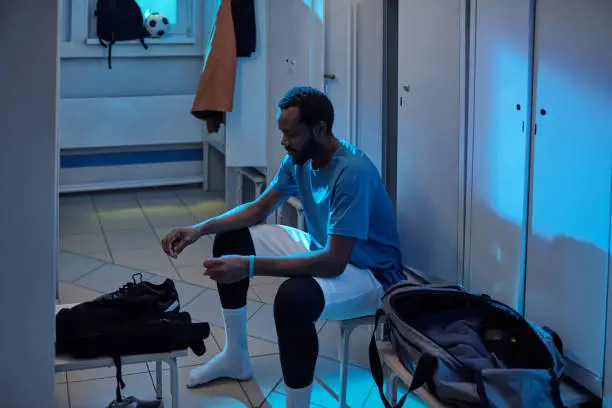 Young African soccer player going to put on his sports-shoes while sitting by row of lockers in changing-room