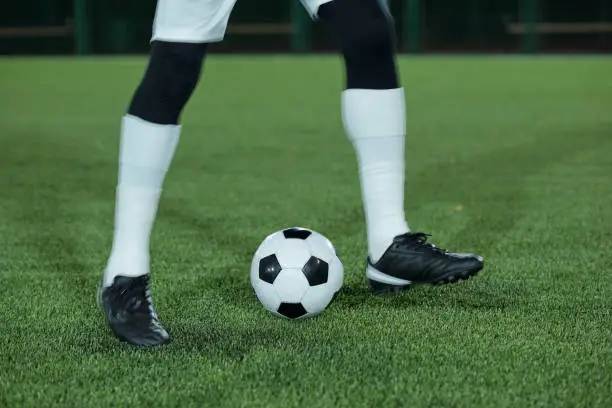 Legs of football player in white socks and black sports-shoes exercising with soccer ball on green lawn at stadium
