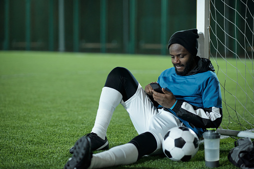 Young restful sportsman sitting by net and scrolling in smartphone while looking through news or online videos