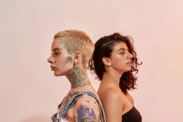 Portrait of two young diverse women, generation Z lesbian couple standing back to back, posing together isolated over light pink background. Lgbtq, youth concept