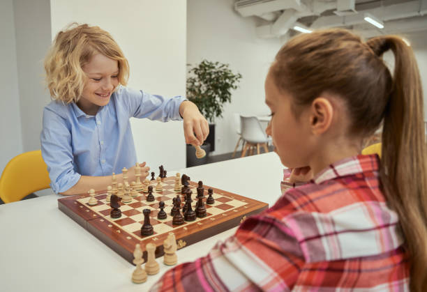 verspielter kleiner kaukasischer junge, der beim zug lächelt, mit einem freund schach spielt, in der schule zusammen am tisch sitzt - concentration chess playing playful stock-fotos und bilder