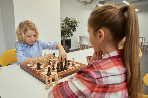 istock Serious little caucasian boy making his move while playing chess with friend, sitting together at the table in school 1354688216