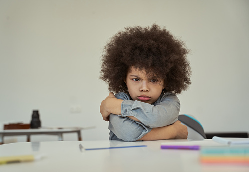 Disgruntled little schoolboy with afro hair looking resentful, frowning while sitting with arms crossed at the table in elementary school classroom