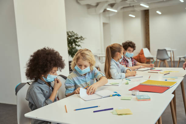istock Little boys wearing protective face mask communicating with each other. Elementary school kids sitting together at the table in a classroom 1354690873