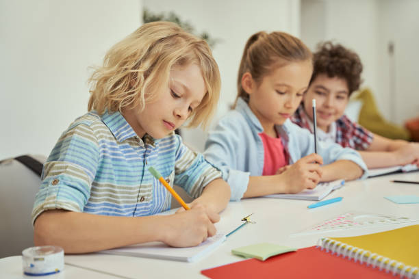 istock Quality education. Diligent schoolboy making notes during lesson. Kids studying, sitting together at the table in elementary school classroom 1354690988