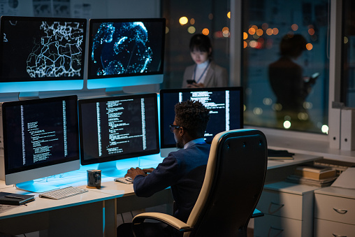 Young African it-developer typing in front of computer screen while decoding data and comparing it with that on another monitor