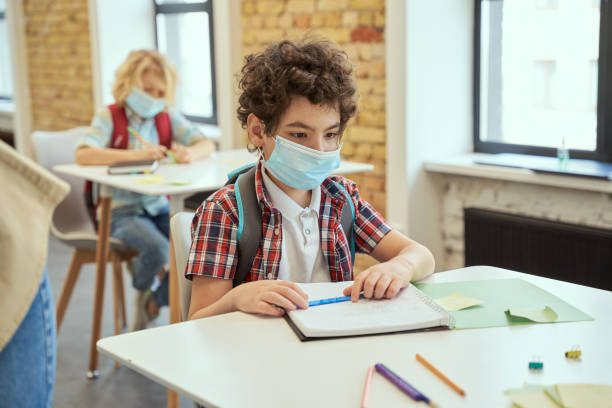 istock Social distancing. School boy wearing face mask writing in his notebook while sitting at the desk in a classroom. Kids studying in elementary school 1354694554