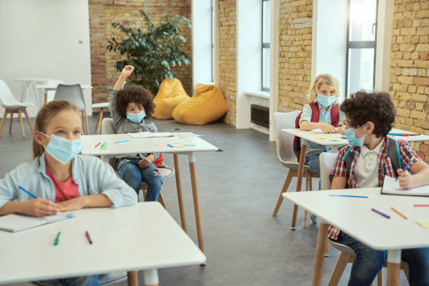 istock Little boy wearing protective face mask during coronavirus outbreak raised his hand in class. School kids studying in elementary school, sitting at the desk 1354694559
